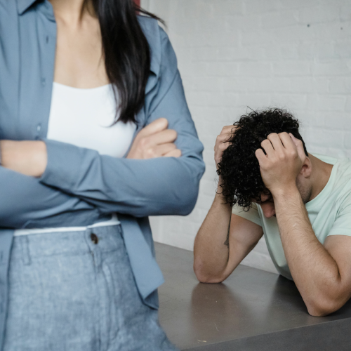 A man sits at a table, heads town, arms to his head. In the foreground, a woman's torso with folded arms faces the camera