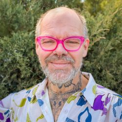 A portait of a man wearing a white patterned shirt, pink glasses, and septum ring standing against natural green backdrop