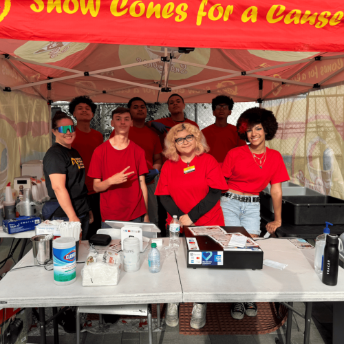 7 youth wearing red shirts and 1 adult woman wearing a black shirt pose under a snow cone canopy