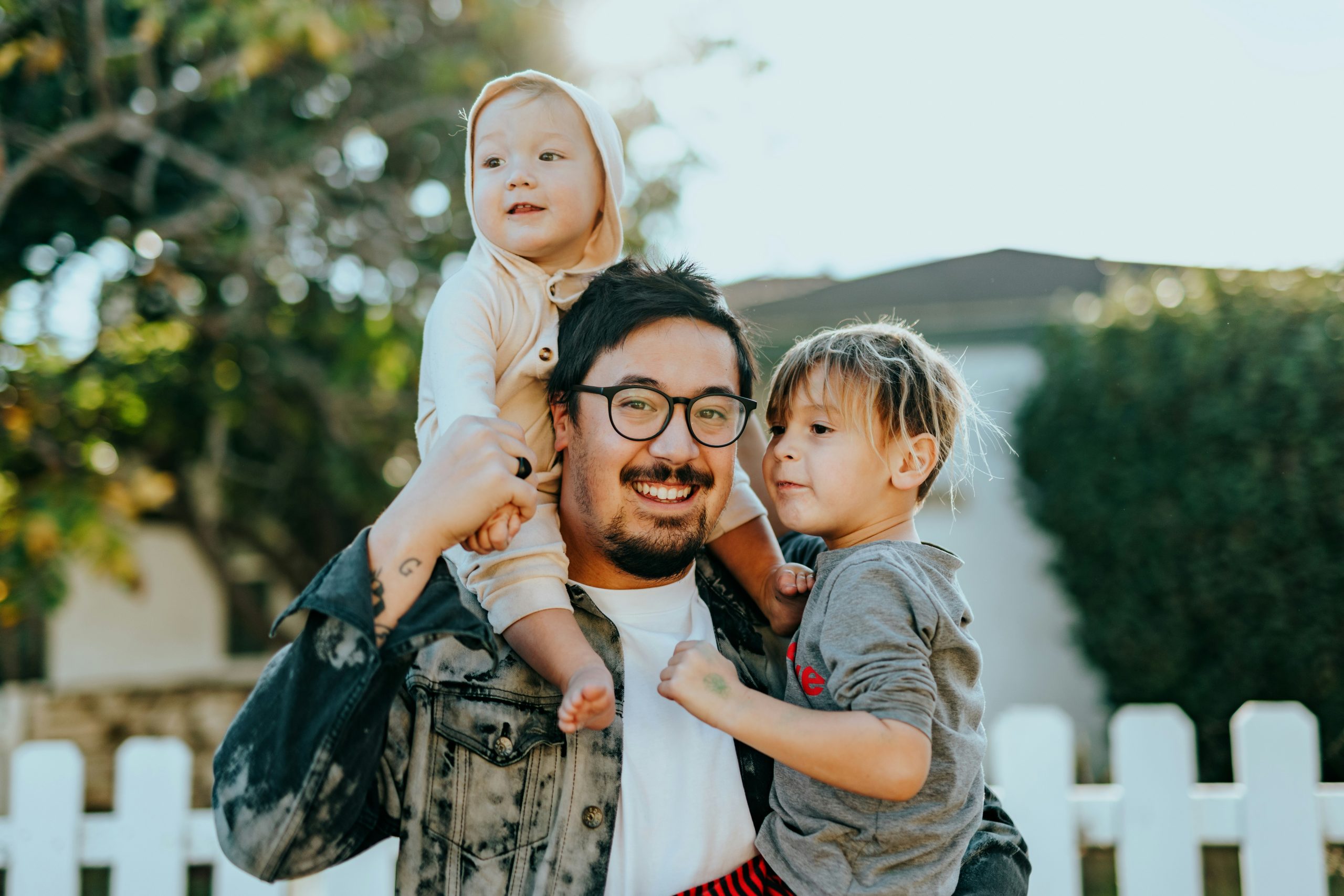 A latino male wearing glasses carries a baby on his shoulders and a small child in the other