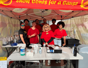 7 youth wearing red shirts and 1 adult woman wearing a black shirt pose under a snow cone canopy